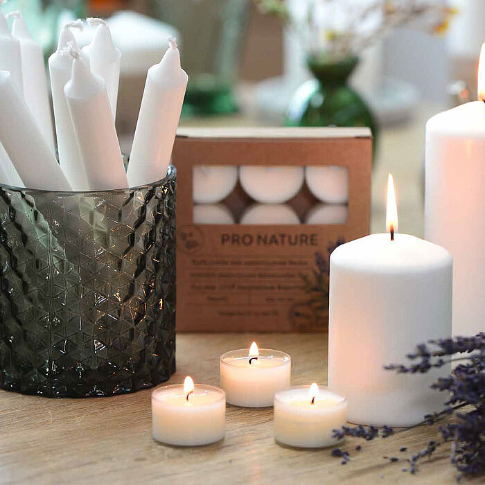 Auf einem Holztisch stehen mehrere brennende weiße Teelichter von Müller. Daneben befinden sich zwei brennende weiße Stumpenkerzen von Müller in unterschiedlichen Größen und ein schwarzes, facettiertes Glas, das mit weißen Stabkerzen von Müller gefüllt is
