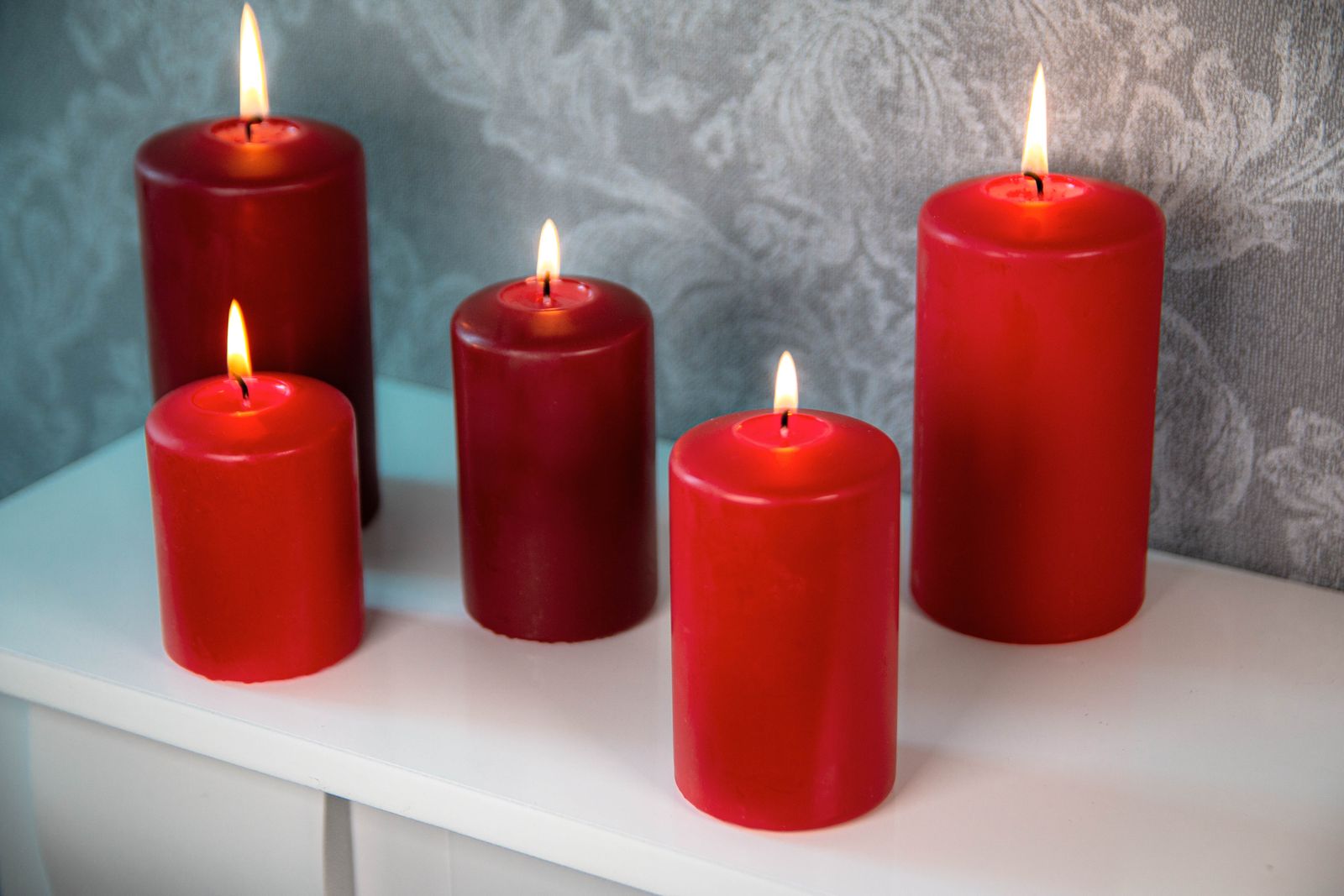 Fünf brennende rote Stumpenkerzen in verschiedenen Größen stehen auf einer weißen Oberfläche vor einem dezent gemusterten grauen Hintergrund. Die Produkte sind von DecoLite.